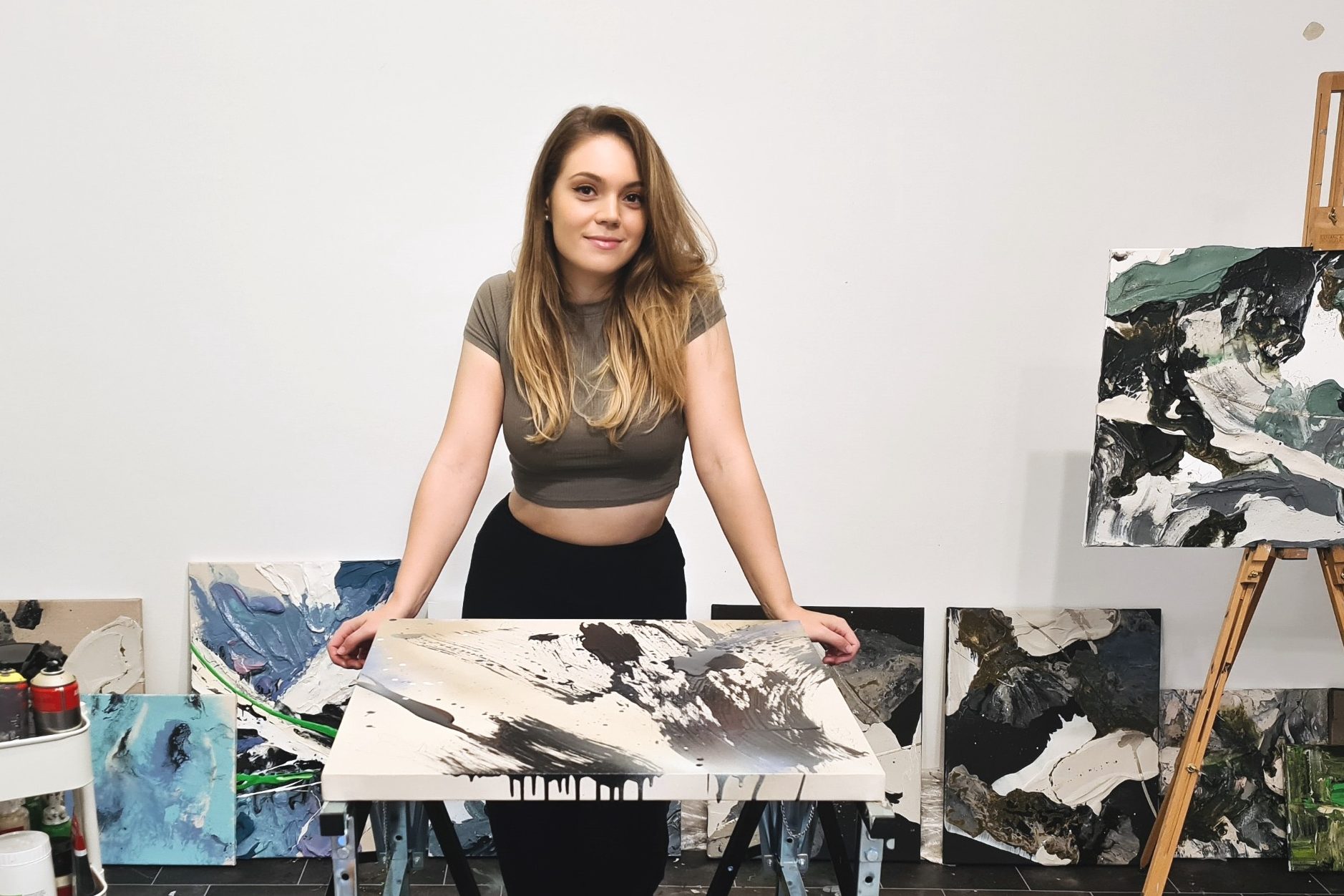 Kseniya Krei Arts Junge Künstlerin steht lächelnd vor ihren abstrakten Gemälden in einem Atelier.
